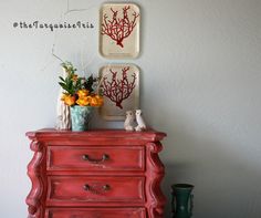 a red dresser with two pictures on the wall above it and vases filled with flowers