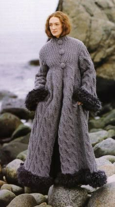 a woman standing on some rocks by the water wearing a gray coat with fur trims