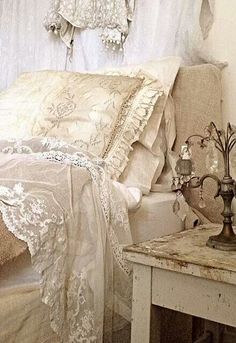 a bed with white sheets and lace on it's headboard, next to a small side table