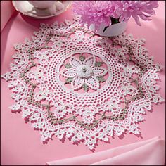 a pink table cloth with white crochet doily and flowers in a vase