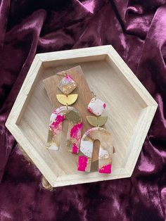 a wooden tray with pink and gold earrings on it, sitting on a purple cloth