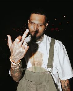 a tattooed man making the vulcan sign with his hand while wearing overalls and a white t - shirt