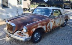 an old rusted car is parked in front of a house with a trailer behind it