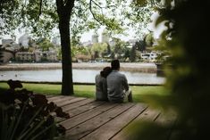 two people sitting on a wooden deck in front of a body of water and trees