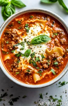 a white bowl filled with pasta and meat soup on top of a table next to basil leaves