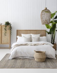 a bed with white linens and pillows in a bedroom next to a basket, plant and wicker cabinet