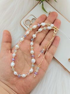 a hand holding a long necklace with pearls and crystals on it, next to a cell phone