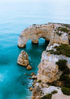 an arch shaped rock formation in the middle of the ocean with blue water and green vegetation