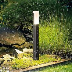 a light that is sitting in the grass near some rocks and plants with rain coming down on it