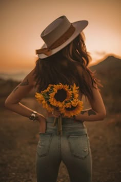 a woman with a hat and sunflowers in her back, looking at the sunset