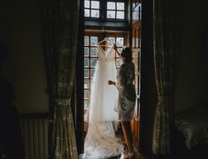 a woman standing in front of a window next to a wedding dress