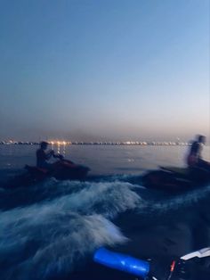 two people riding jet skis on top of a body of water at night with city lights in the background