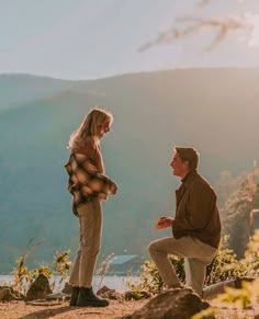 a man kneeling down next to a woman on top of a hill with mountains in the background