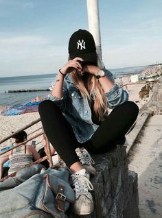 a woman sitting on top of a stone wall next to the ocean wearing a yankees hat