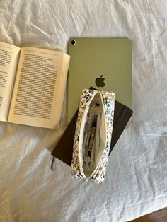 an open book sitting on top of a bed next to an apple laptop and purse