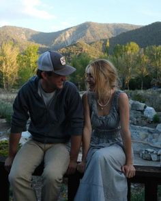 a man sitting next to a woman on top of a wooden bench in front of mountains