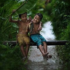 two children sitting on a bench in the rain with an umbrella over their head and one holding a fish