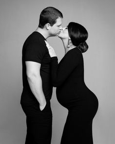 a pregnant couple kissing each other while standing next to each other in black and white