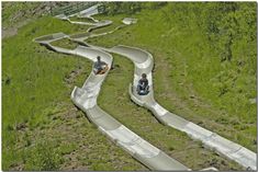 two people riding down a slide in the middle of a grassy area with trees on both sides