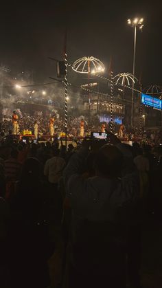 the crowd is watching performers on their cell phones at night in front of an outdoor stage with umbrellas and lights