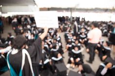 blurry image of graduates in gowns and caps at graduation ceremony with one holding up a sign