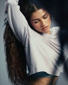 a woman with long hair wearing a white shirt and black shorts is holding her arms behind her head