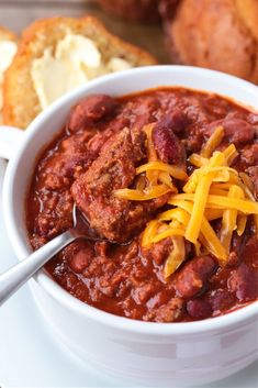 a white bowl filled with chili and cheese on top of a table next to bread