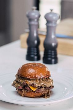 a hamburger sitting on top of a white plate next to two pepper shakers in the background