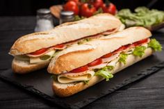 two sub sandwiches with lettuce and tomato on a black slate platter next to tomatoes