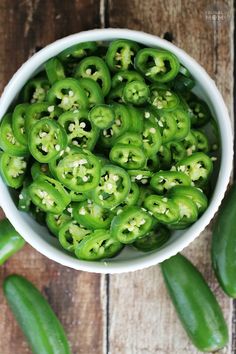 a white bowl filled with green peppers sitting on top of a wooden table next to sliced jalapenos