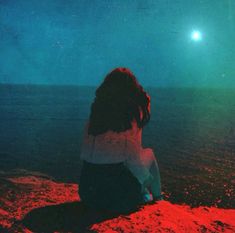 a woman sitting on top of a beach next to the ocean under a full moon