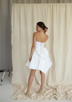 a woman standing in front of a backdrop wearing a white dress with bow at the waist