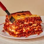 a piece of lasagna on a white plate with a red handled spatula