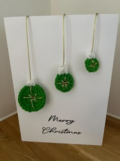 three green crocheted christmas ornaments hang from gold - plated hooks on a white card