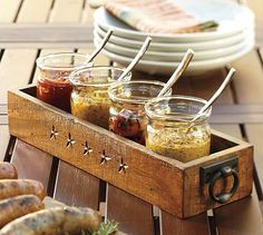 four jars of food sit in a wooden tray on a table with plates and utensils