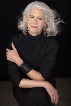 an older woman with white hair and black dress posing for a photo in front of a dark background