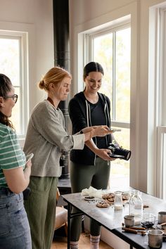 three women standing in front of a table with food on it and one woman holding a camera