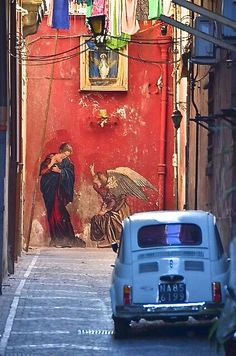 an old car parked on the side of a street next to a wall with paintings