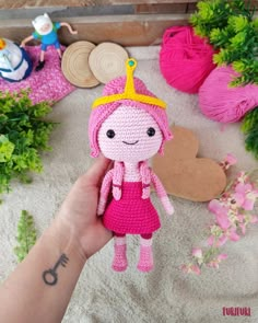 a hand holding a pink crocheted doll in front of some flowers and yarn