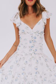 This exclusive Wisteria Lane dress is a playful and stylish addition to any wardrobe. The cream base is embellished with a delicate light blue floral print and embroidered details. The midi length and flutter sleeves add a touch of whimsy, while the decorative buttons, ruching, and ruffled tiers provide a flirty and feminine touch. Perfect for a day out in the sun or a special occasion. Details Cream dress with light blue floral print Cream embroidered details Nonfunctional buttons down front of Spring Floral Dress With Ruffle Sleeves For Brunch, Feminine Floral Dress With Ditsy Print And Flutter Sleeves, Summer Floral Dress With Flutter Sleeves, Feminine Ditsy Floral Print Dress With Flutter Sleeves, Feminine Floral Dress With Ruffle Sleeves For Spring, Summer Floral Dress With Ruffle Sleeves For Brunch, Feminine Floral Dress With Flutter Sleeves For Brunch, White Flutter Sleeve Dress With Ditsy Floral Print, Spring Floral Dress With Flutter Sleeves For Garden Party