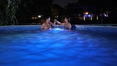 two people sitting in the middle of a swimming pool at night, one holding a camera