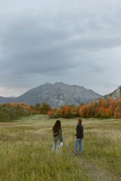 two people are walking through the grass in front of mountains and trees with orange leaves