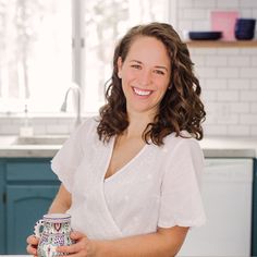 a woman holding a coffee mug in her hand and smiling at the camera while standing next to a kitchen counter