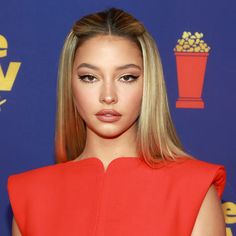 a woman with long blonde hair wearing a red dress and looking at the camera while standing in front of a blue backdrop