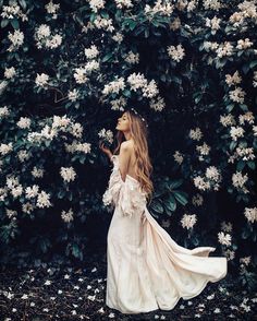 a woman in a white dress standing next to a bush with flowers on the wall