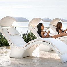 two beautiful women sitting on lounge chairs next to each other in front of the ocean