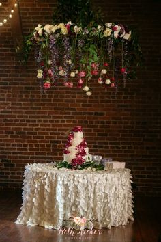 a wedding cake sitting on top of a table next to a brick wall with flowers hanging from it