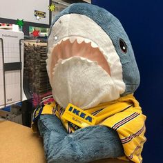 a stuffed shark with a yellow shirt on it's chest and mouth is sitting in front of a computer desk