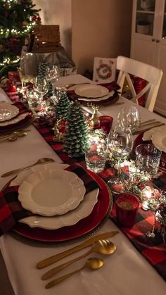 the table is set for christmas dinner with red and white plates, silverware, and evergreen trees