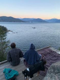 two people sitting on a dock with a dog looking out at the water and mountains in the distance